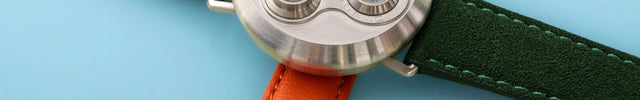 Drei Uhren mit grünen und orangefarbenen Uhrenarmbändern auf hellblauem Hintergrund